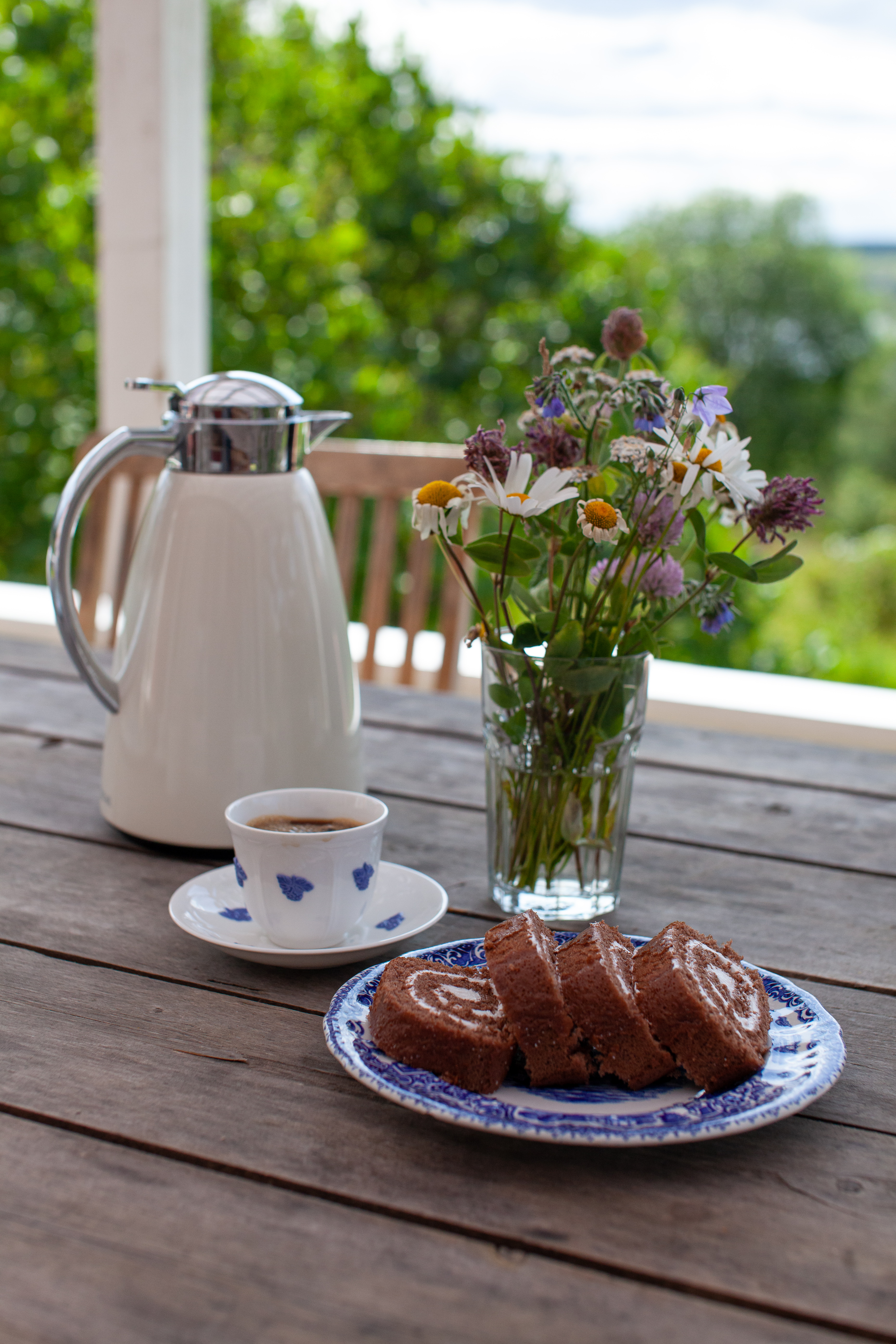 Vit kaffetermos, blåvit kaffekopp med fat, ett fat med rulltårteskivor och en vas med sommarblommor.