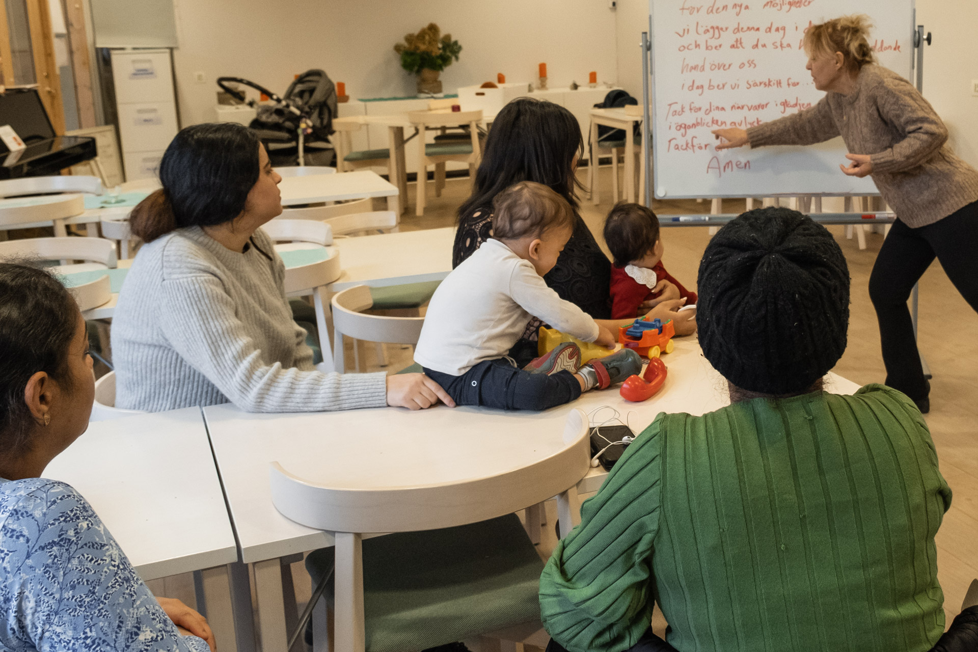 Fyra kvinnor och två barn sitter vid ett bord. De tittar mot en kvinna som står upp och pekar mot en text på en whiteboardtavla