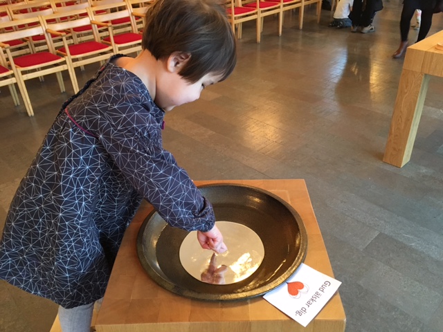 Barn står vid en dopfunt och doppar sina fingrar i vattnet. Vid dopfunten ligger en lapp där det står "Gud älskar dig".