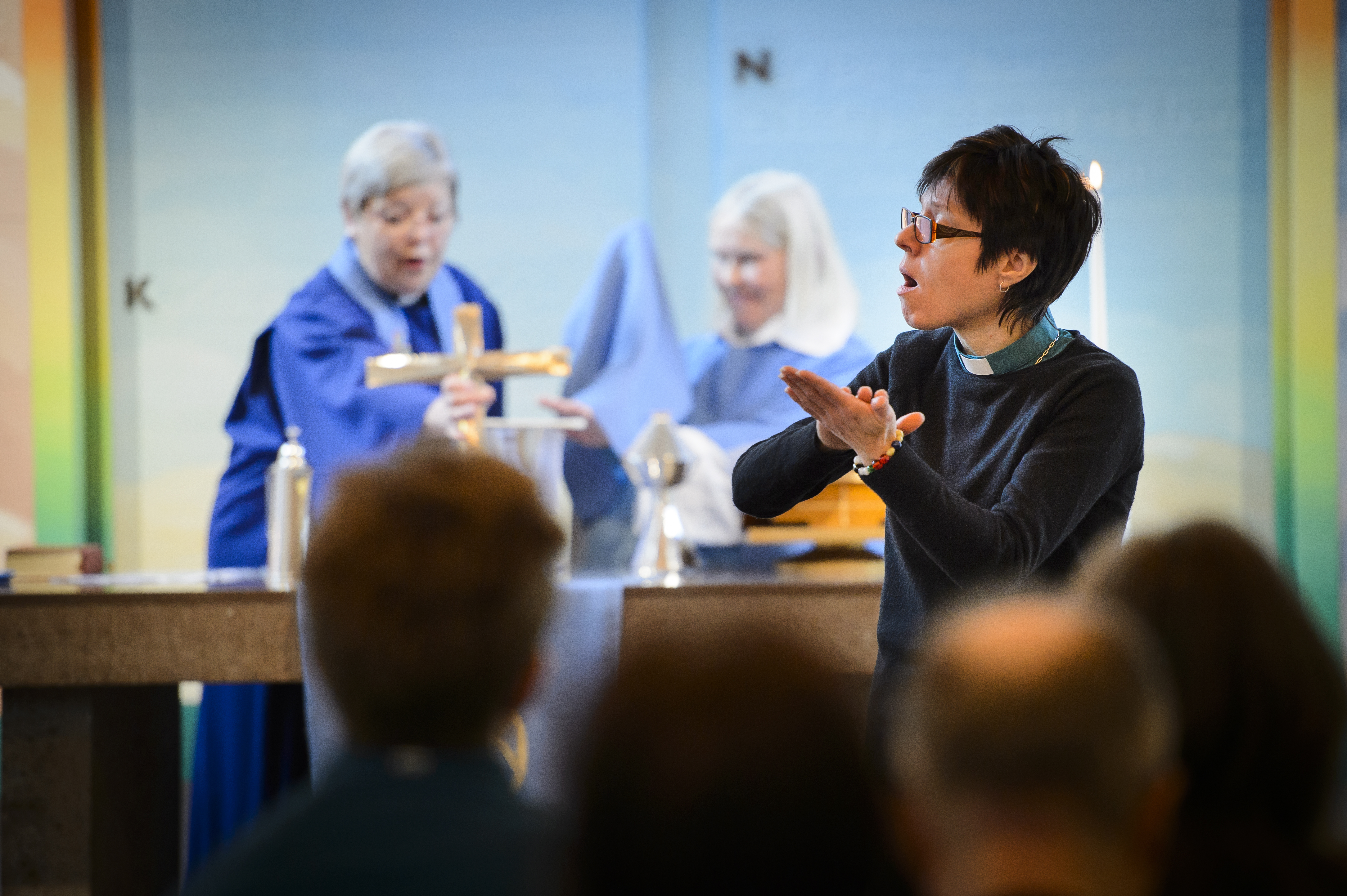 En kvinnlig präst talar teckenspråk. Några besökare i förgrunden och två kvinnliga präster i bakgrunden.