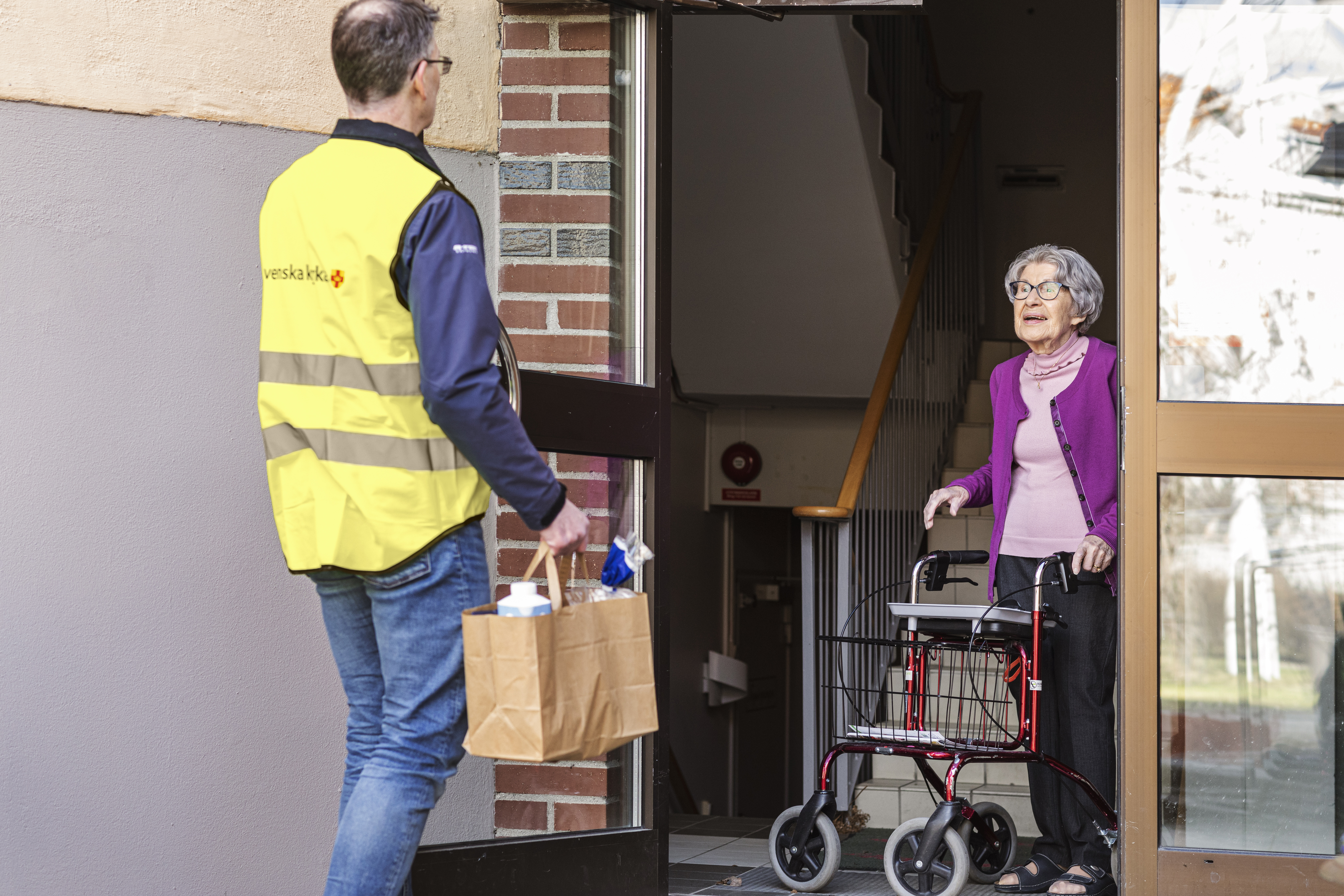 En man i gul reflexväst med Svenska kyrkan logotype överräcker en matkasse till en äldre kvinna med rollator.