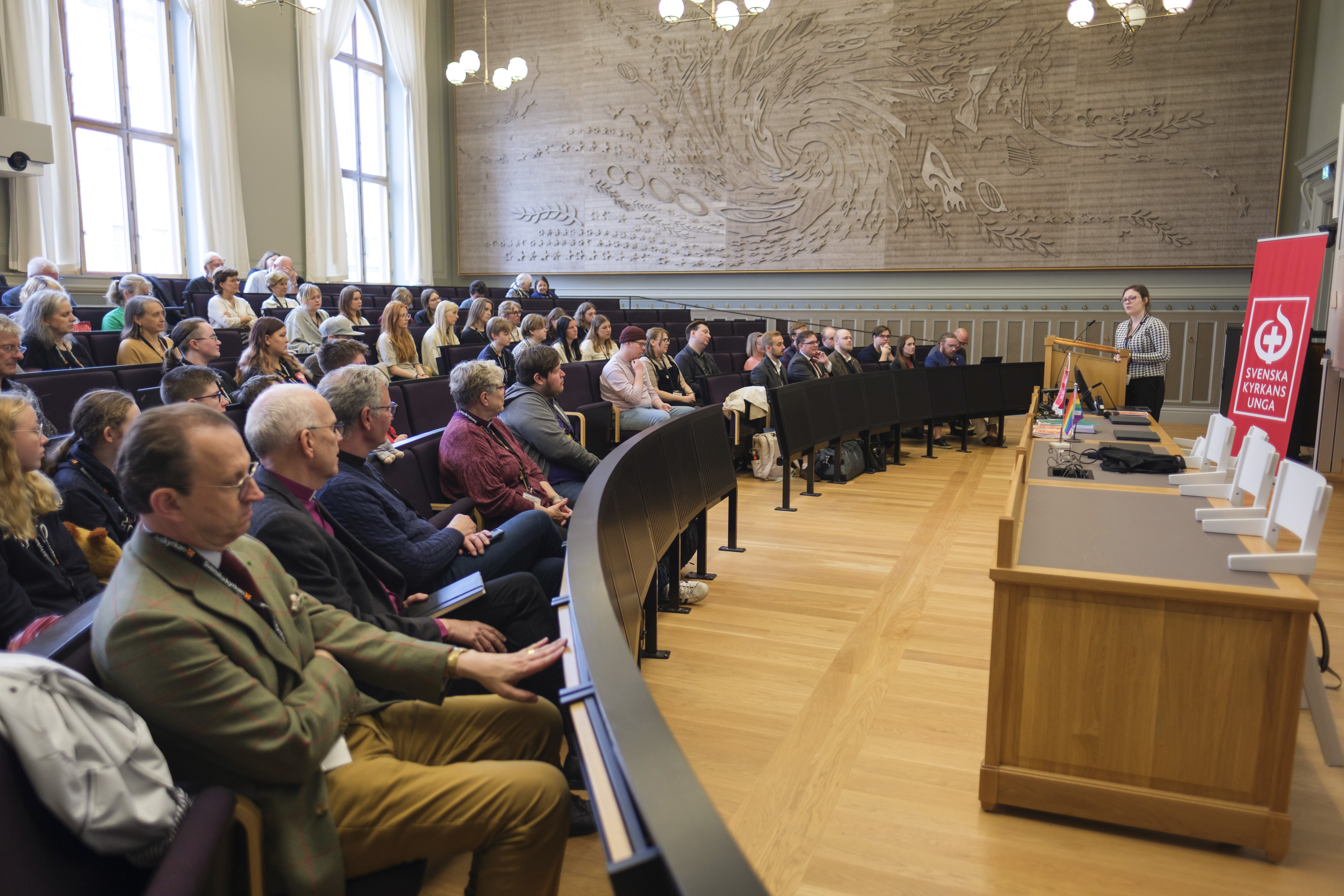 En kvinna föreläser för en nästan fullsatt sal. På en röd rollup står texten Svenska kyrkans unga.