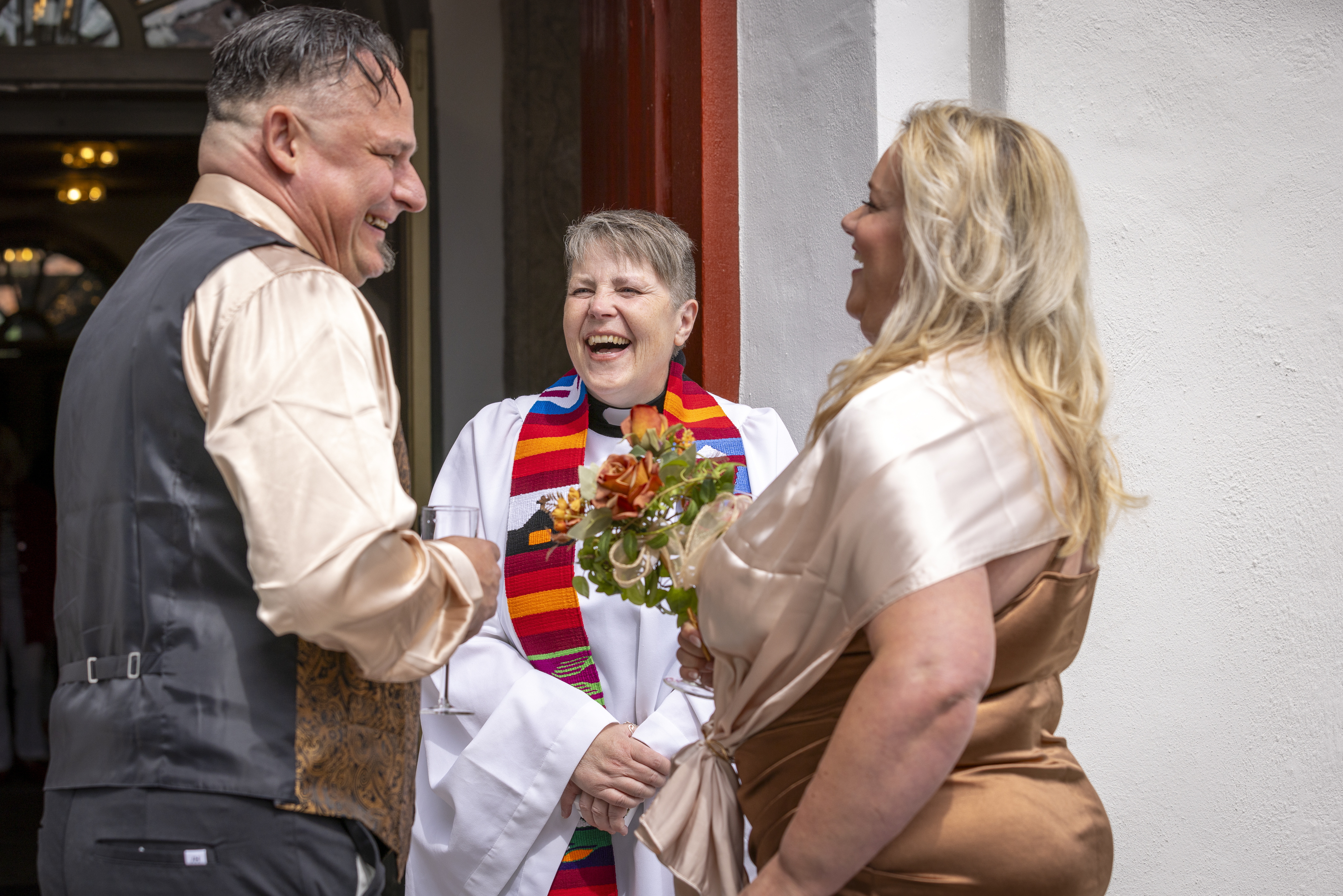Drop-in vigsel. Ett brudpar står vid kyrkporten tillsammans med en präst och skrattar. Bruden håller en blombukett i handen.
