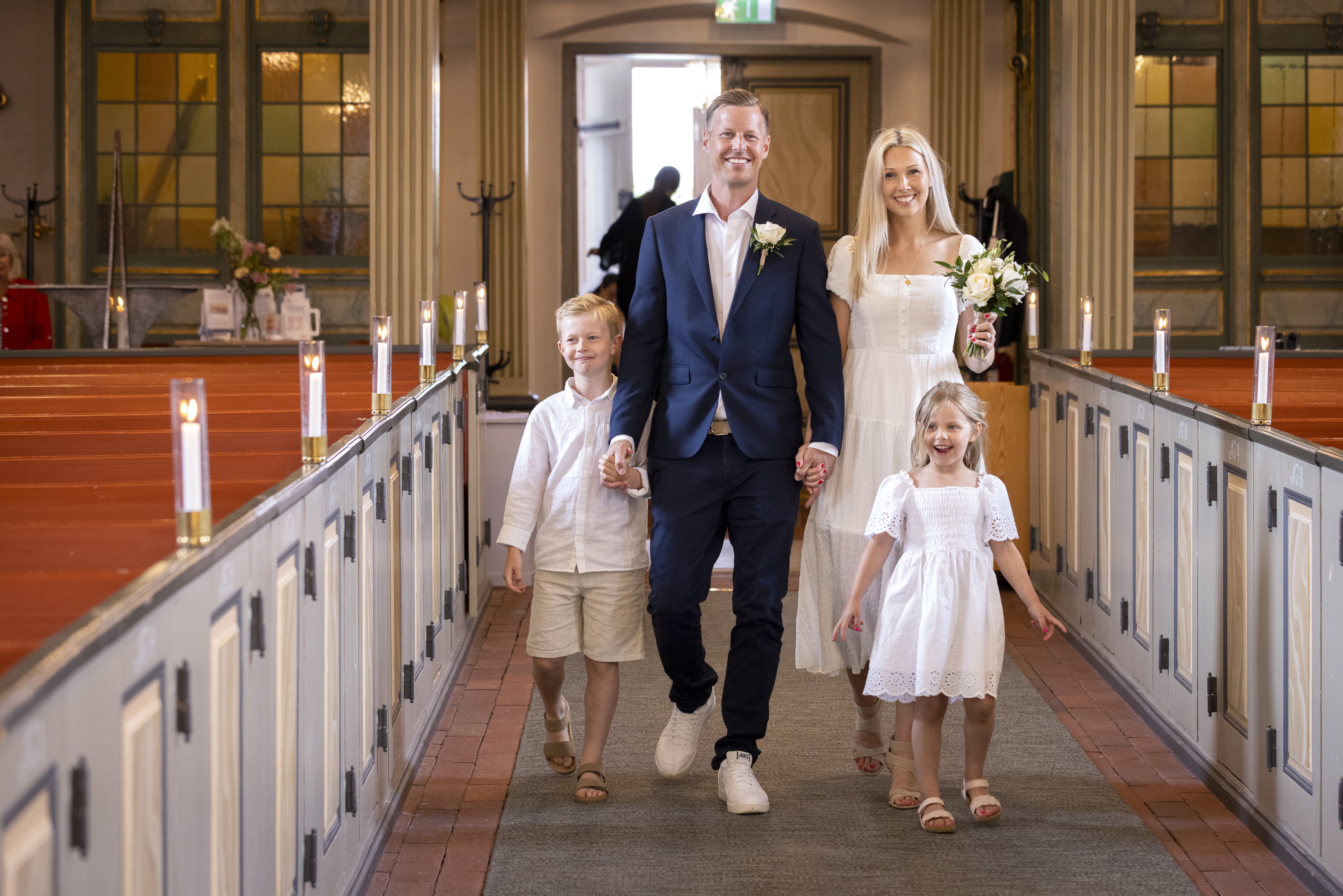 Drop-in vigsel i en kyrka. Ett brudpar går i altargången tillsammans med två ljusklädda barn. Bruden håller en vit blombukett i handen.