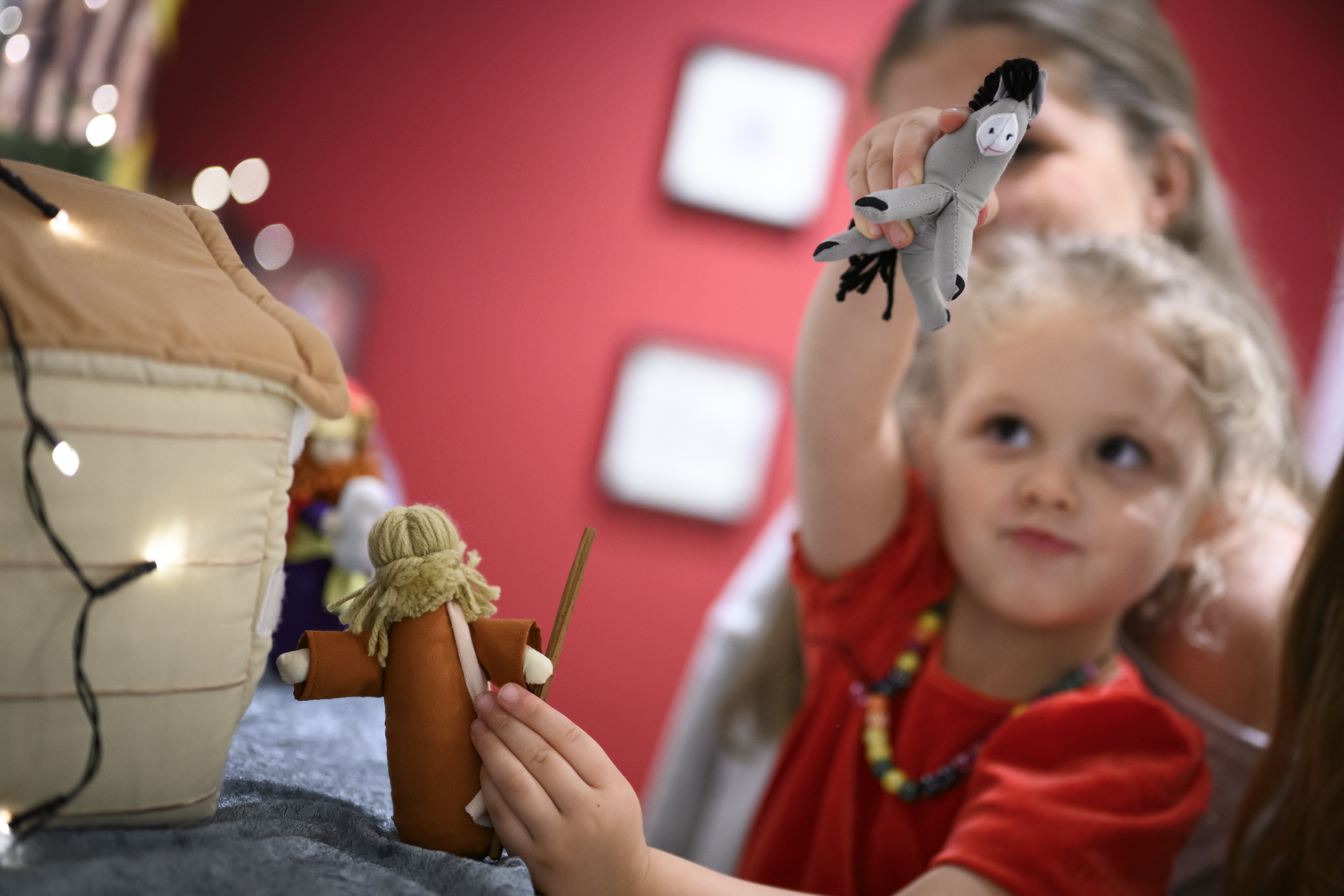 Ett litet barn sträcker med ena handen upp en mjukisåsna mot kameran. En ljusslinga hänger på en julkrubba i mjukistyg.