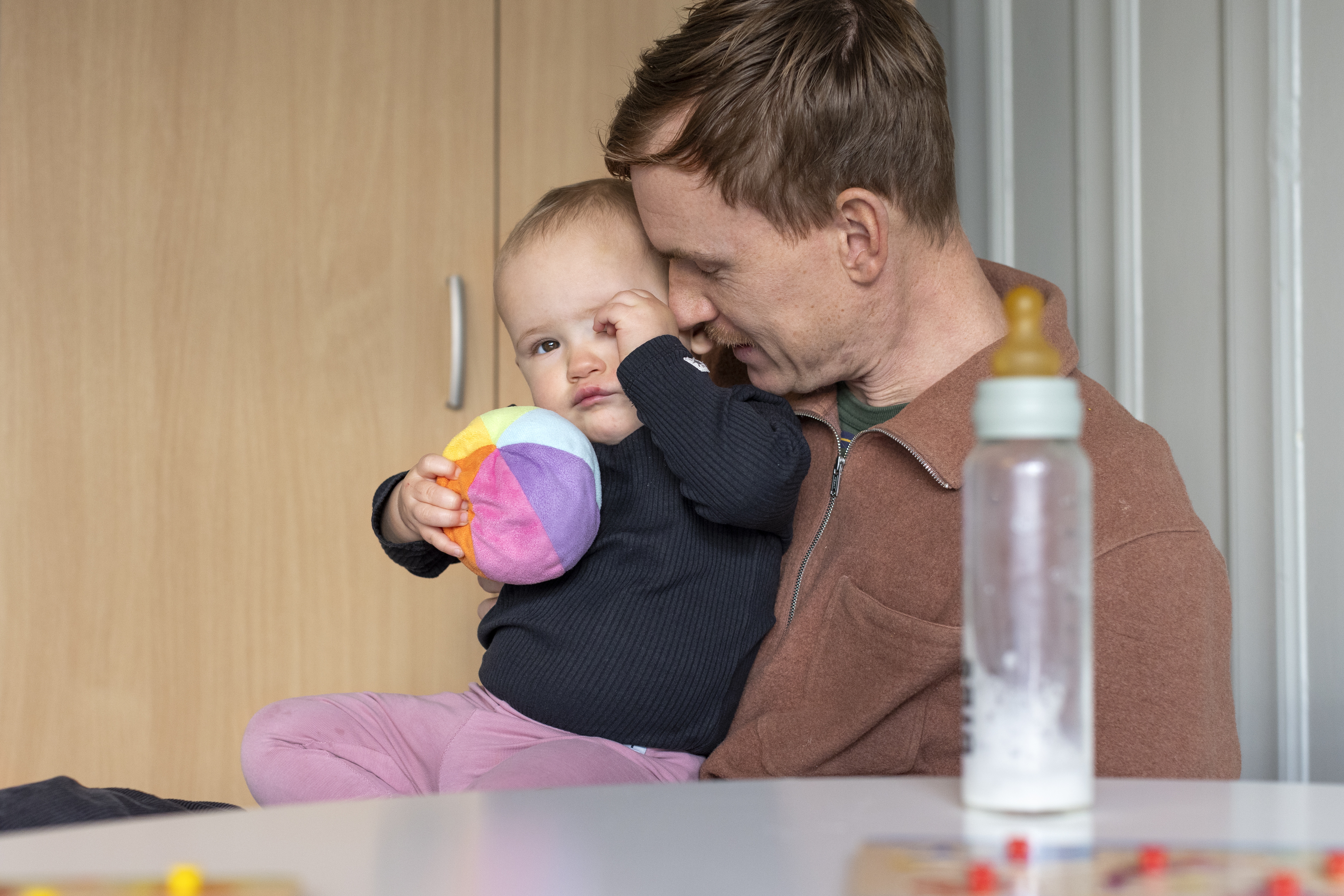 En man sitter med ett litet barn i famnen. Barnet har en färgglad mjuk boll i ena handen. En nappflaska står på bordet.