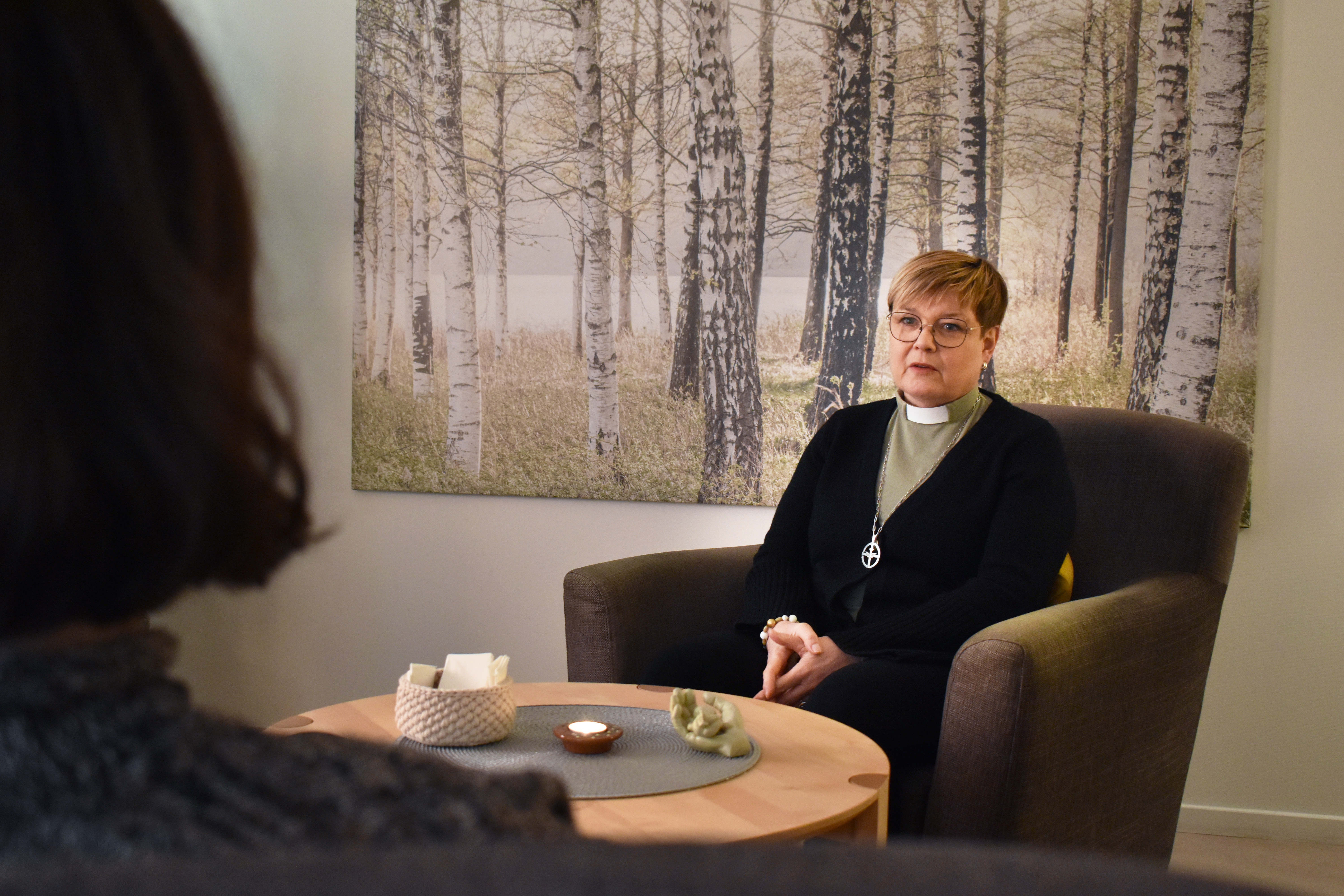 Kvinna med diakonskjorta sitter i fåtölj och samtalar med någon.